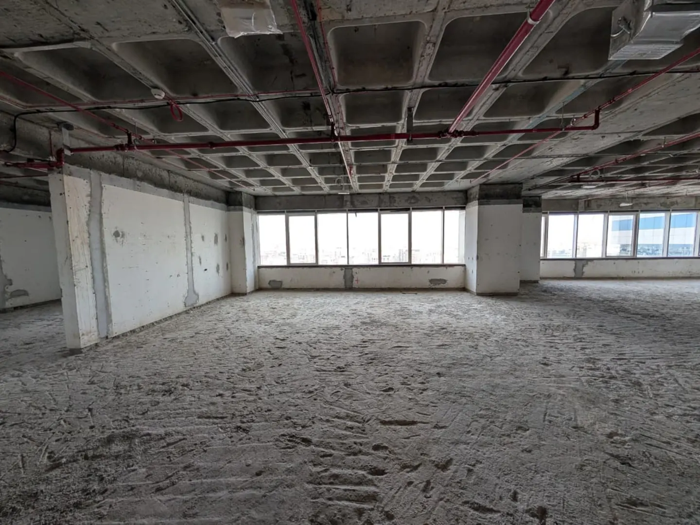 Unfinished office space with concrete floors and ceiling. Red pipes run along the ceiling. Windows line the far wall.