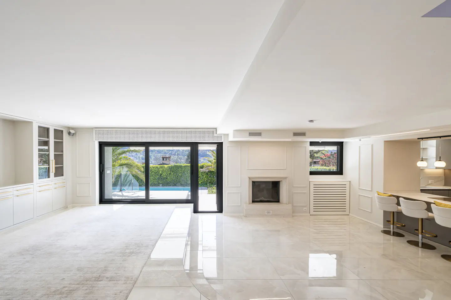 Bright, modern living room with white tile floors, fireplace, and bar seating. Sliding glass doors open to a pool and green landscape.