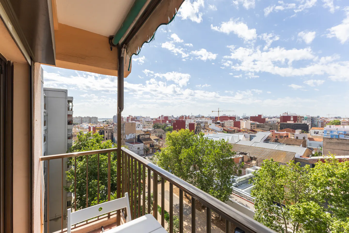 Balcony view of a city with trees, buildings, and a blue sky with white clouds. Brown metal railing and white table and chairs are visible.