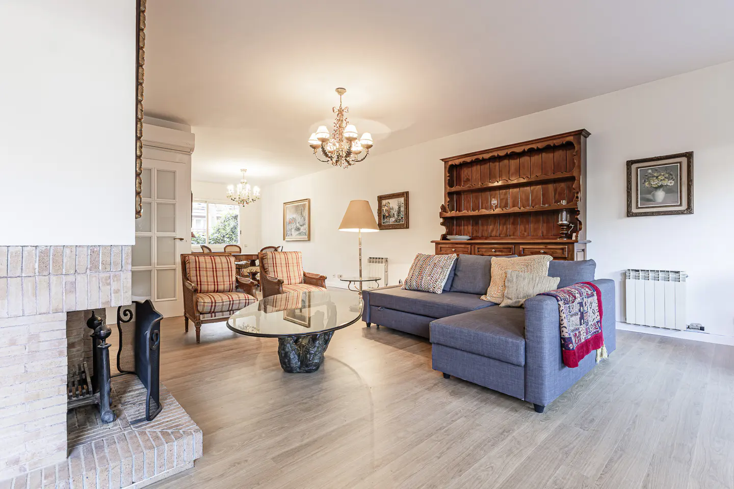 Bright living room with a blue sofa, wooden hutch, and a glass coffee table. A brick fireplace is on the left.