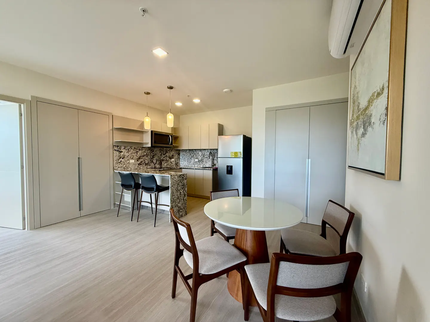 A modern apartment interior with a round dining table, chairs, and a kitchen with granite countertops and stainless steel appliances.