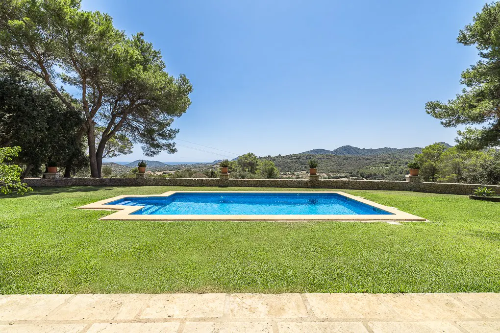 Outdoor pool with blue water surrounded by green grass and trees under a clear blue sky. Stone wall with potted plants in the background.