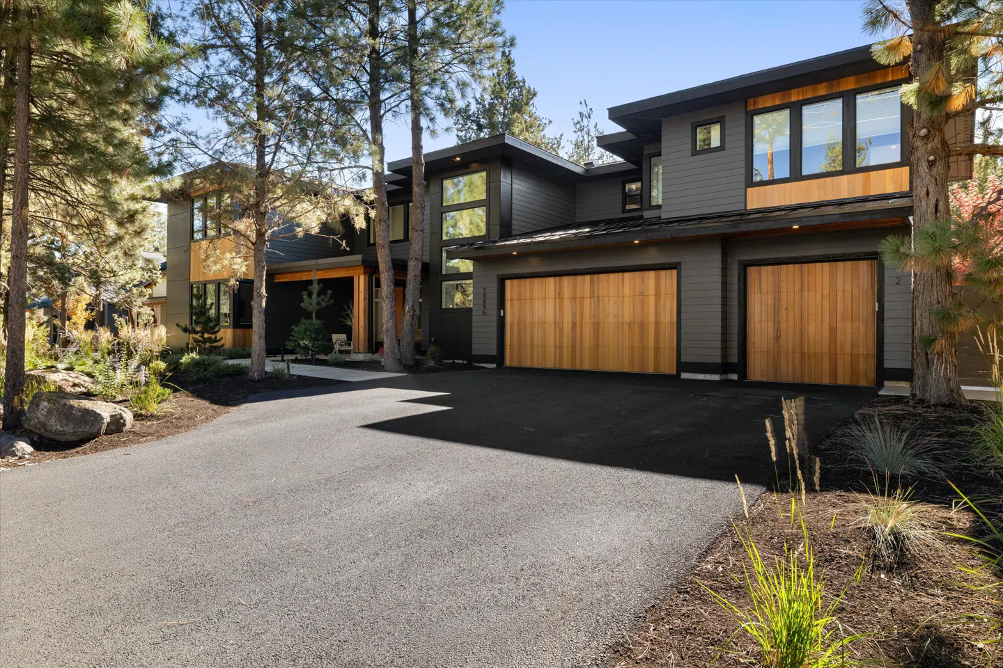 Modern two-story house with gray siding and two wooden garage doors. Tall pine trees surround the house. Black asphalt driveway.