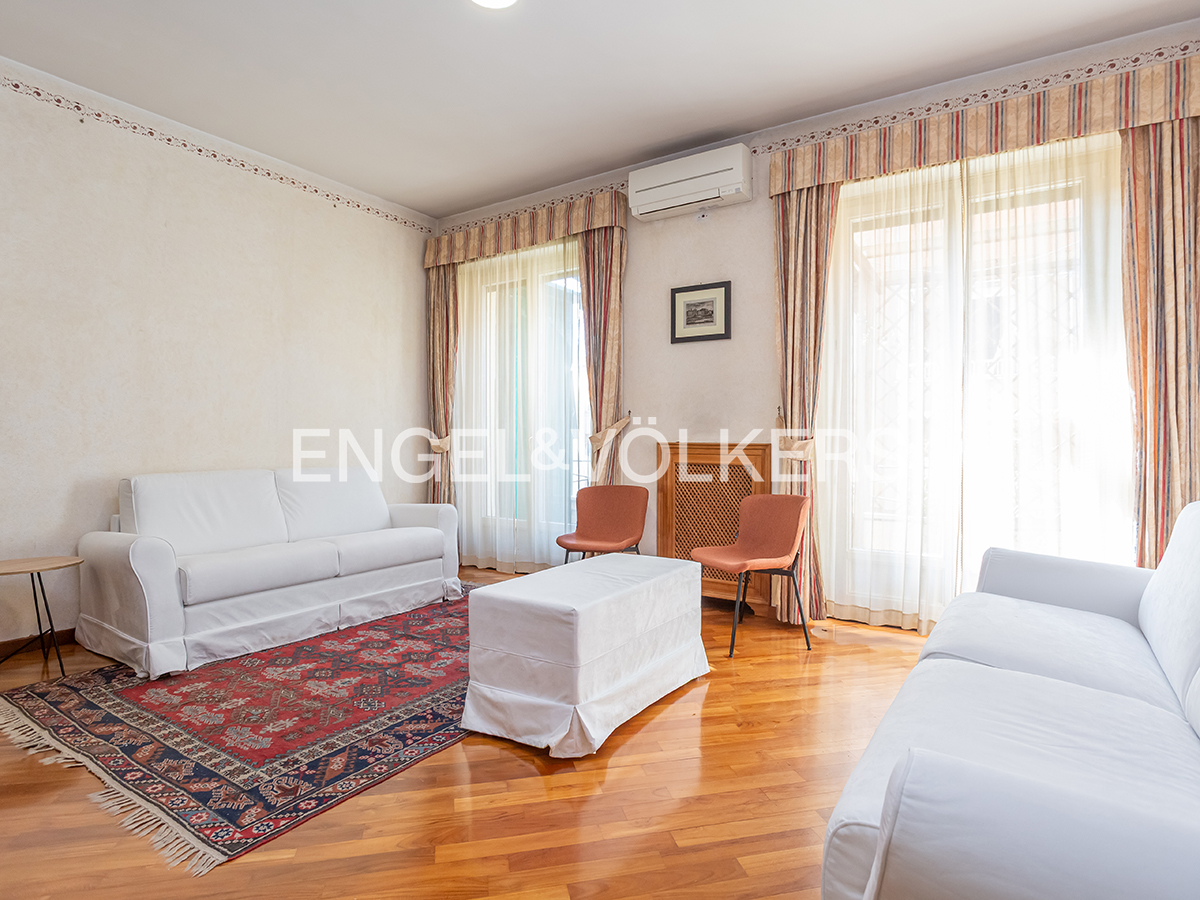 Living room with white sofas, a red patterned rug, and two orange chairs on a wood floor. Curtains frame the windows.