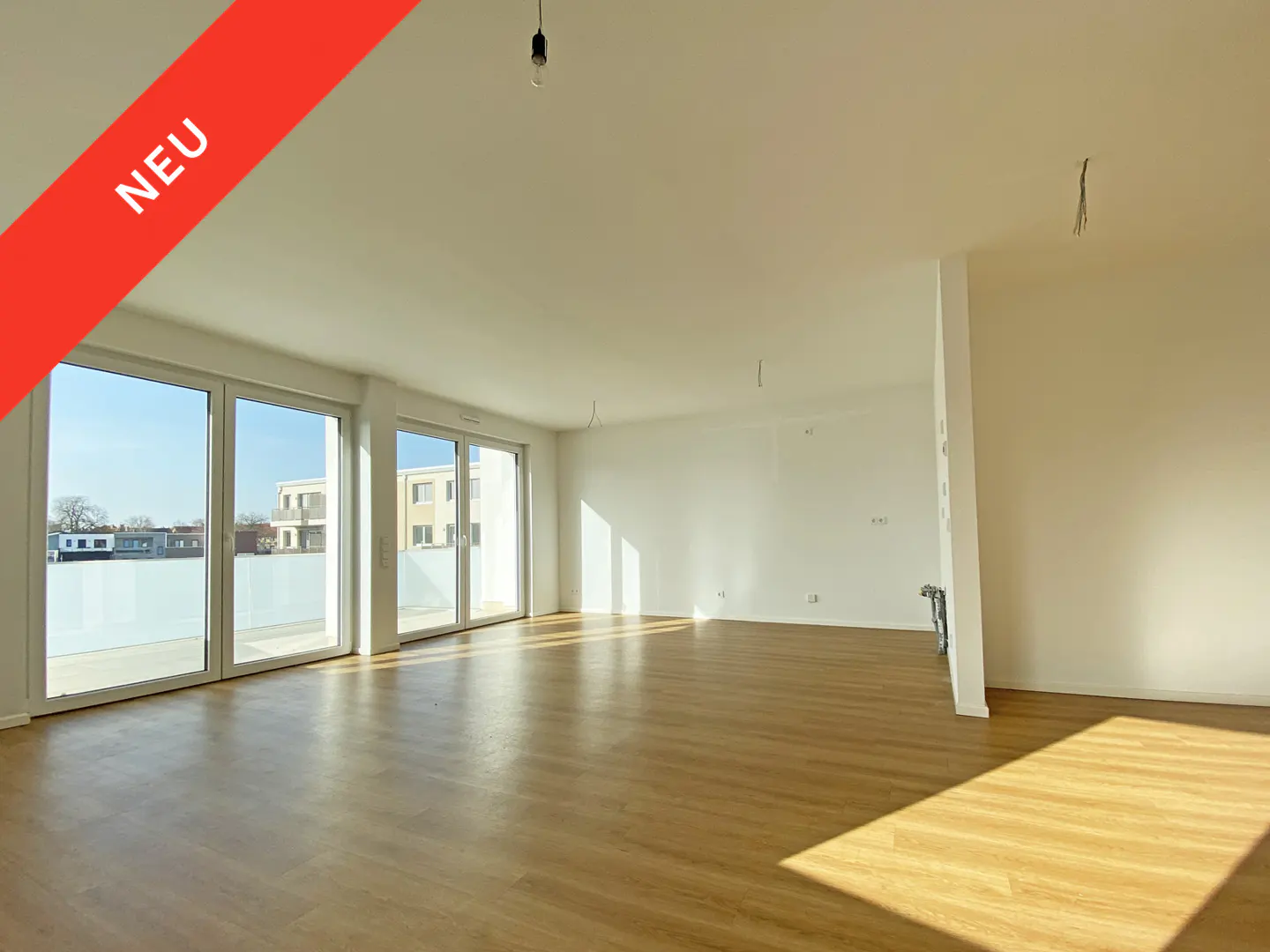 Bright, empty room with wood floors and white walls. Large sliding glass doors lead to a balcony. Red banner with "NEU" text.