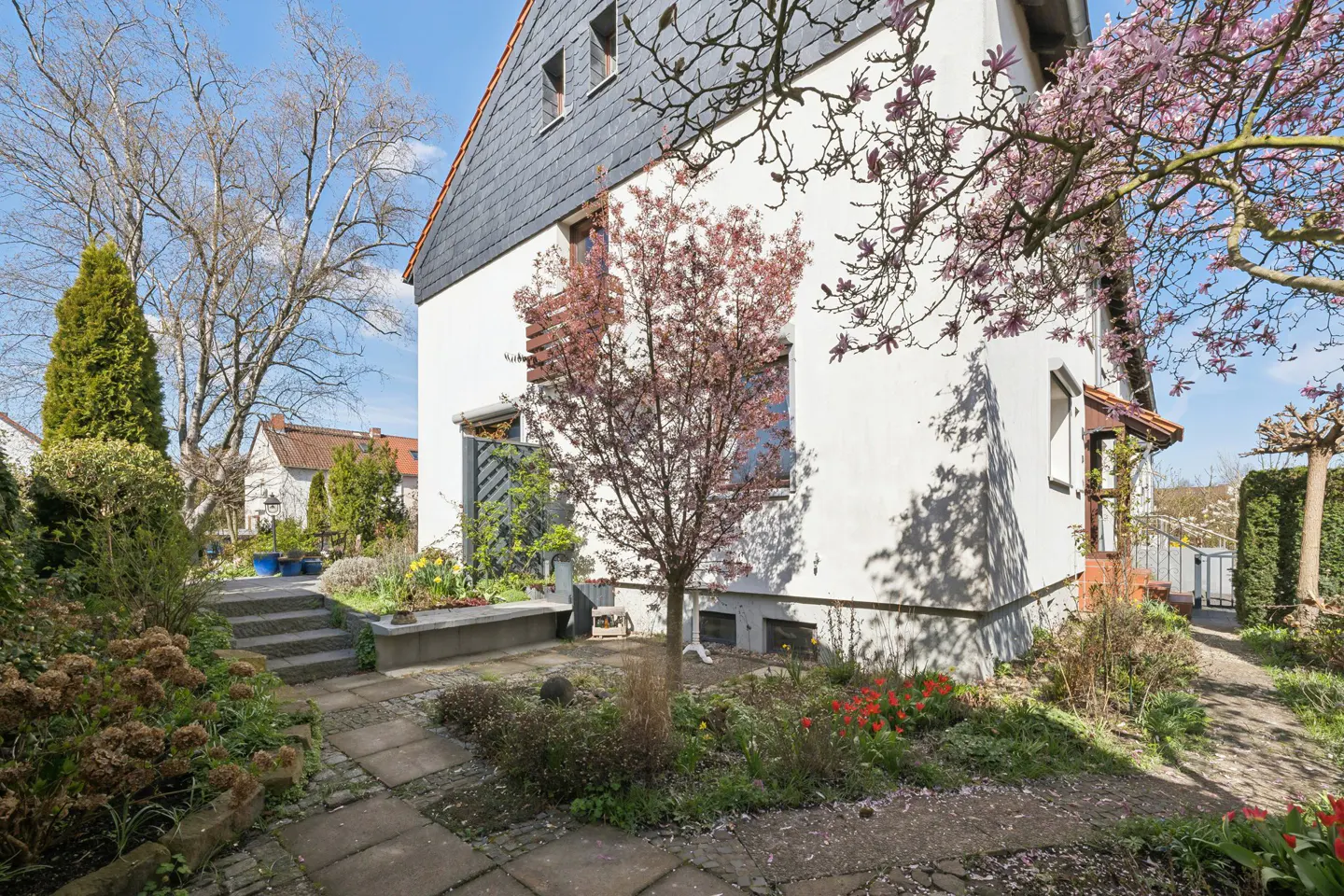 Exterior view of a white house with a gray roof, surrounded by trees and a garden with red flowers.