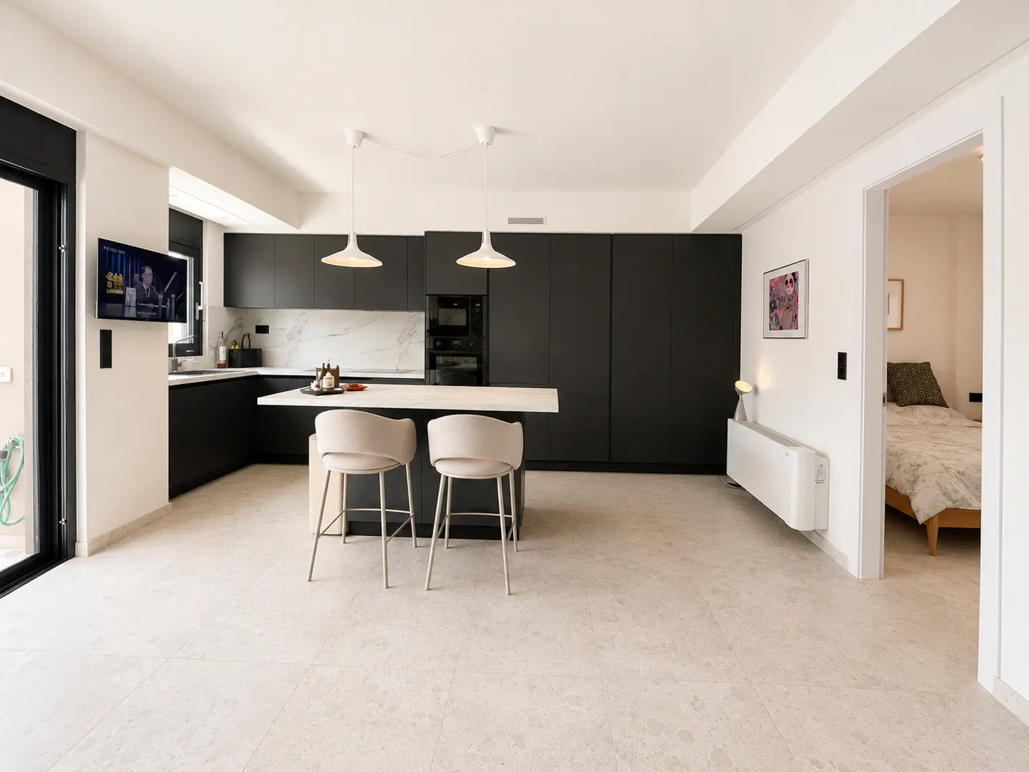 Modern kitchen with black cabinets, white marble backsplash, island with two stools, and a glimpse of a bedroom through a doorway.