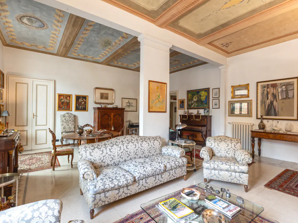 A living room with a floral sofa, armchair, glass table, and a painted ceiling. There are paintings on the walls and a piano in the background.