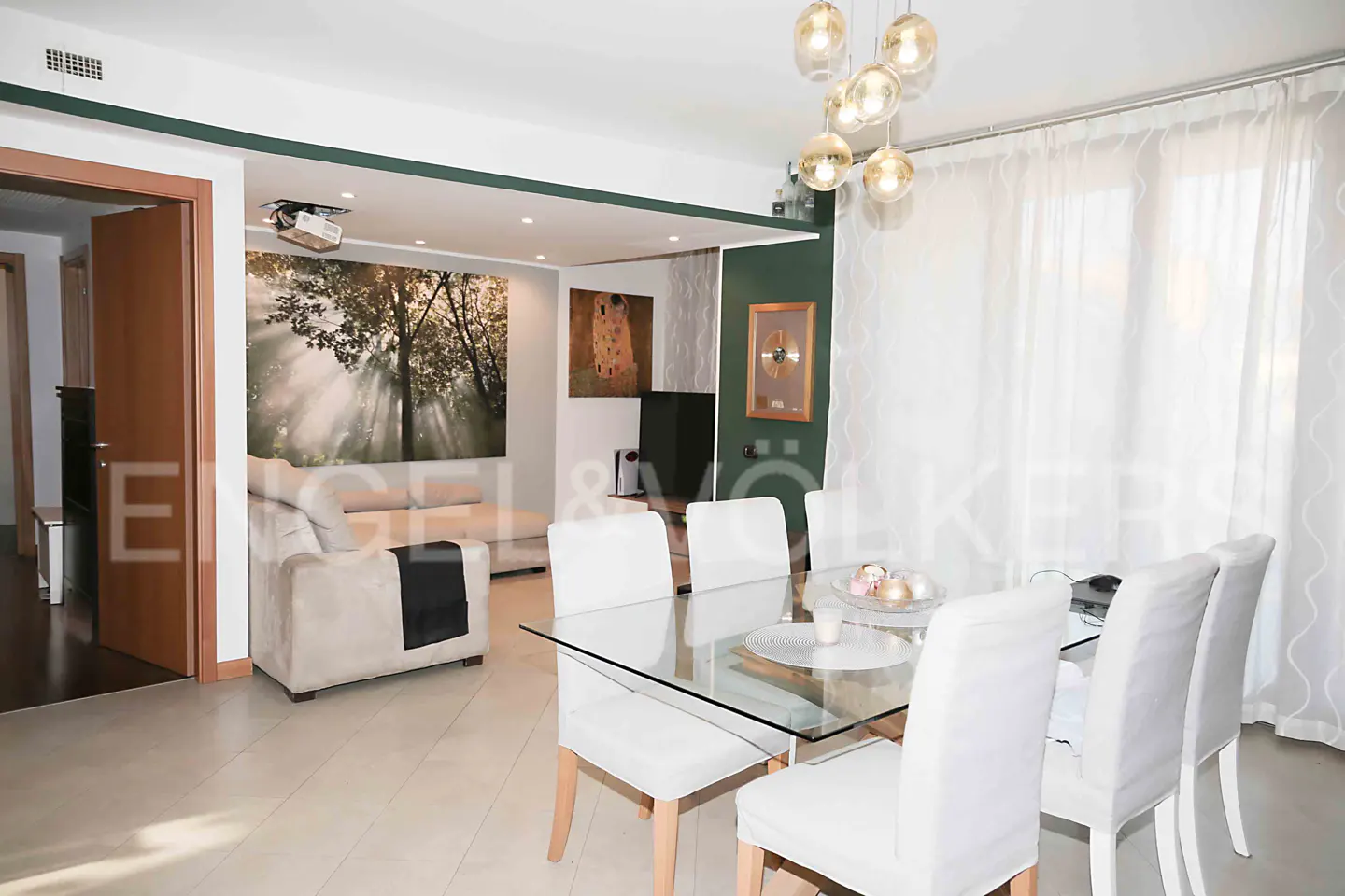 Bright, modern living room with a glass table, white chairs, and a beige sofa. Artwork adorns the walls, and sheer curtains filter natural light.