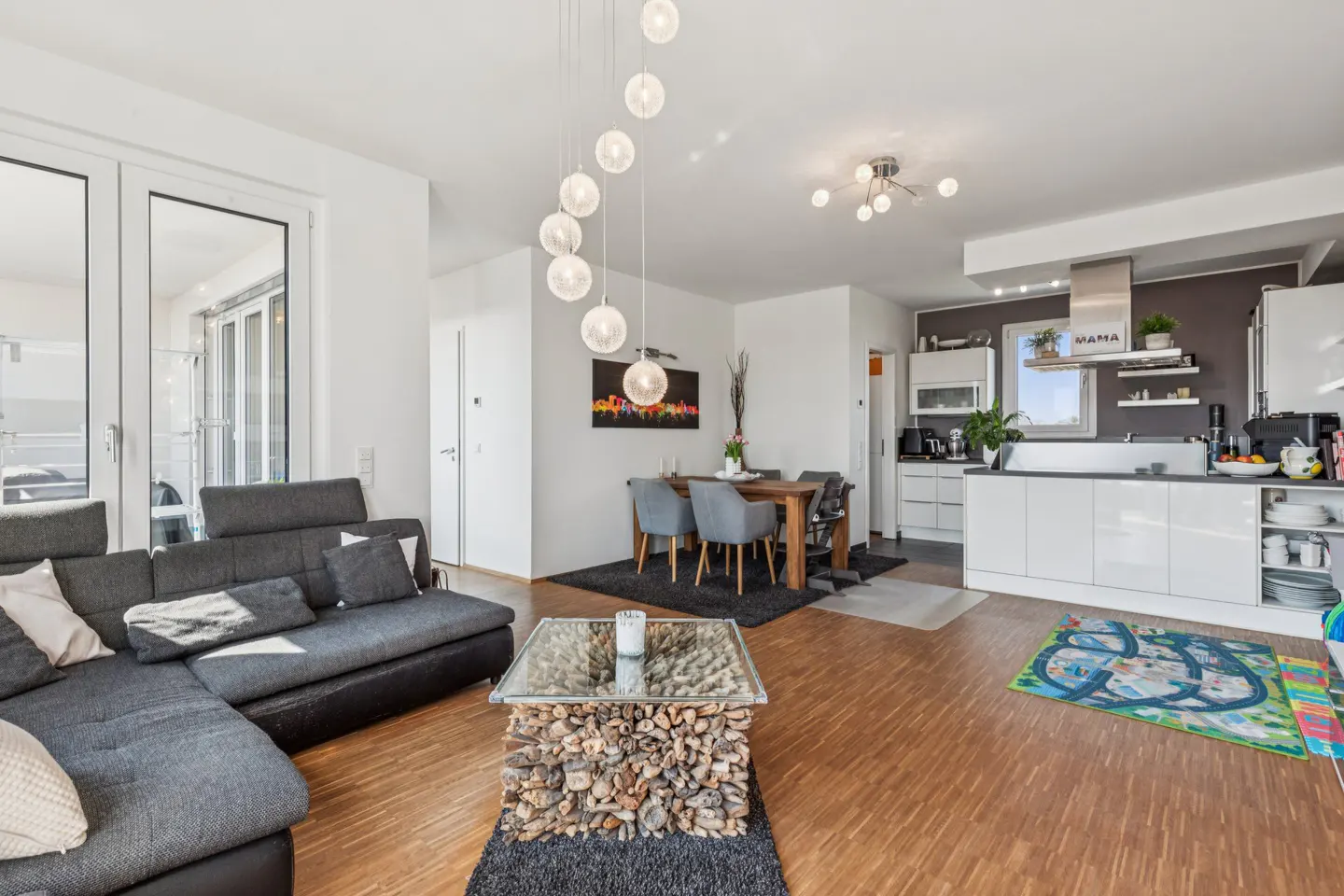 Bright, open-concept living space with gray sectional, wood floors, dining table, and modern white kitchen. A unique coffee table adds character.