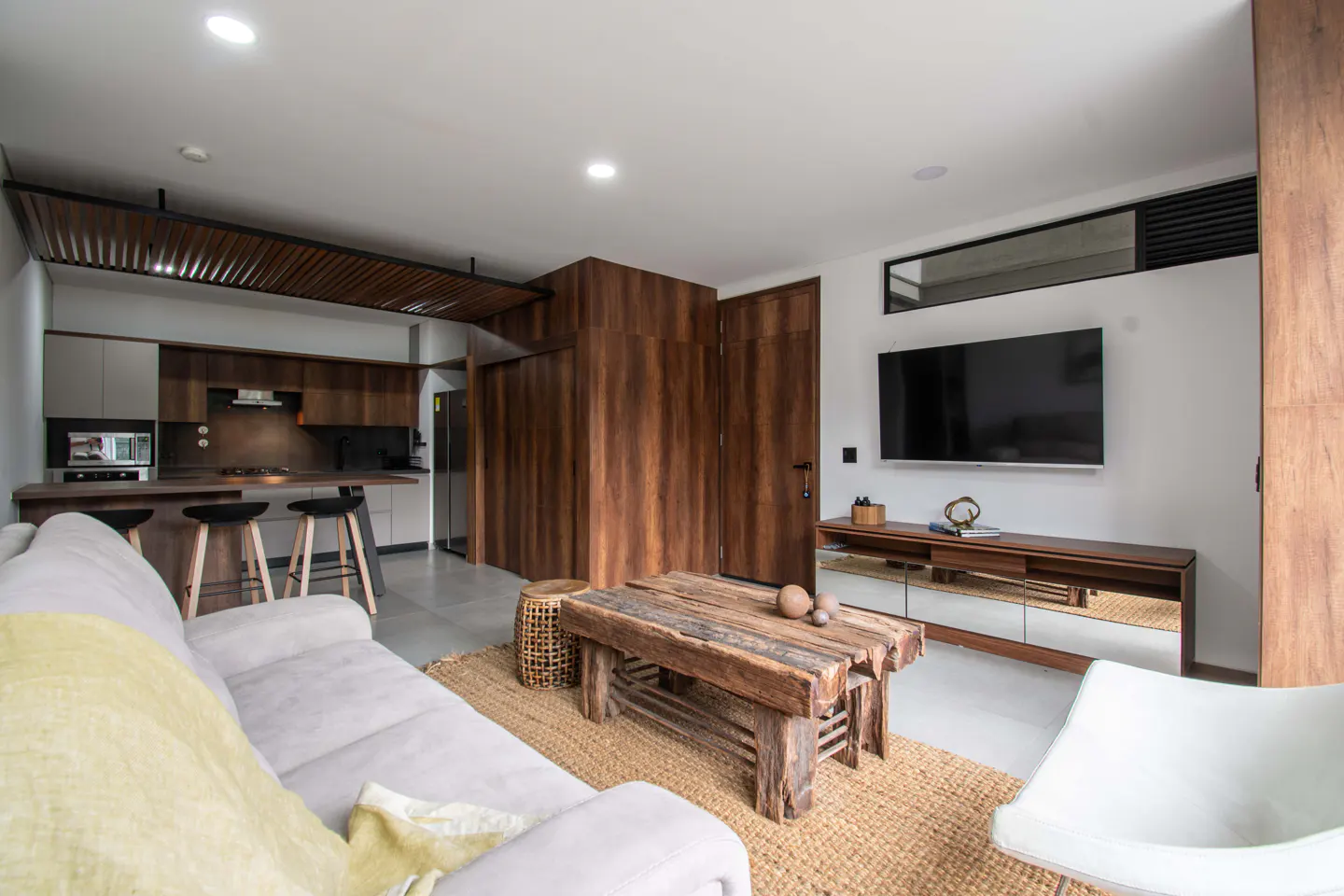Modern living room with a gray sofa, rustic wood coffee table on a jute rug, and a TV mounted on the wall. The kitchen is in the background.