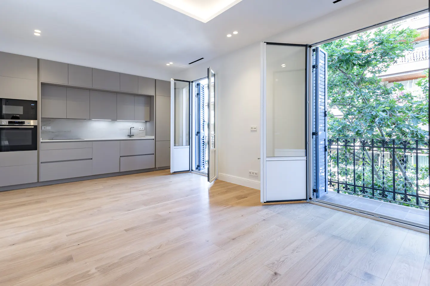 Bright apartment interior with gray kitchen cabinets, light wood floors, and open glass doors to a balcony with trees.
