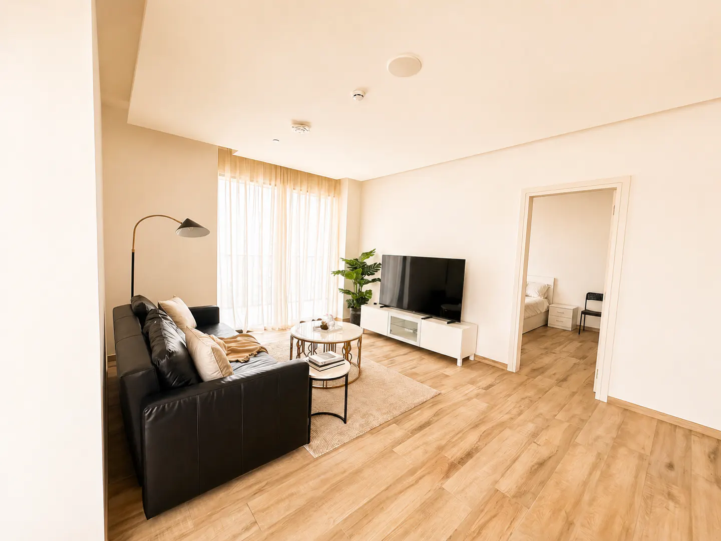 Bright living room with wood floors, black leather sofa, TV, and a view into a bedroom.