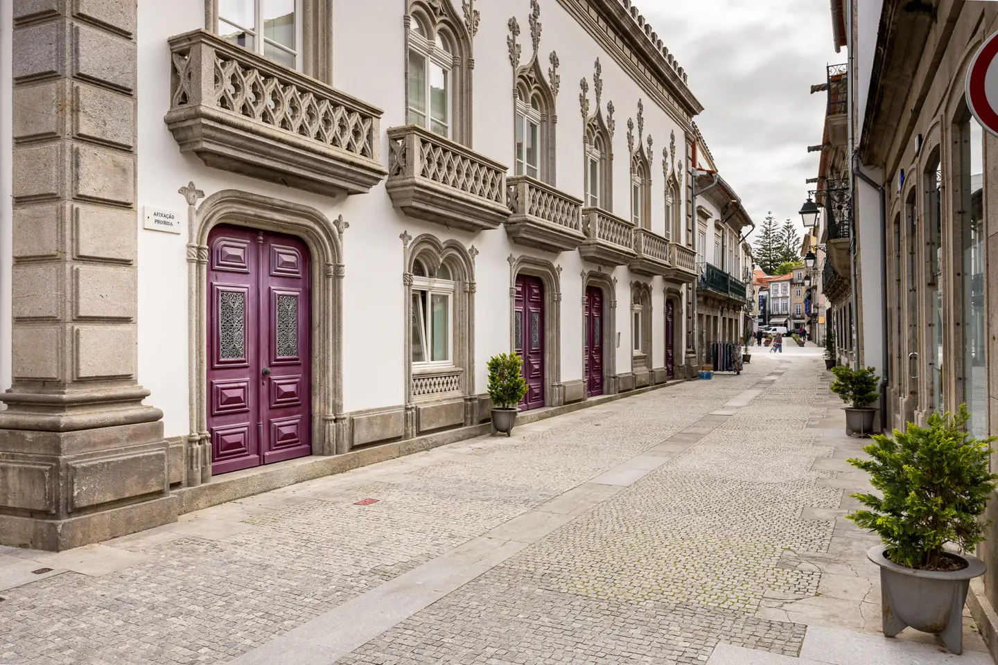 Cobblestone street view with white buildings, purple doors, and stone balconies. Cloudy sky.