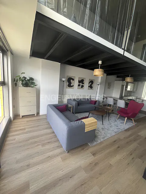 Modern loft living room with grey sofas, a red armchair, wooden floors, and a black industrial ceiling with pendant lights.