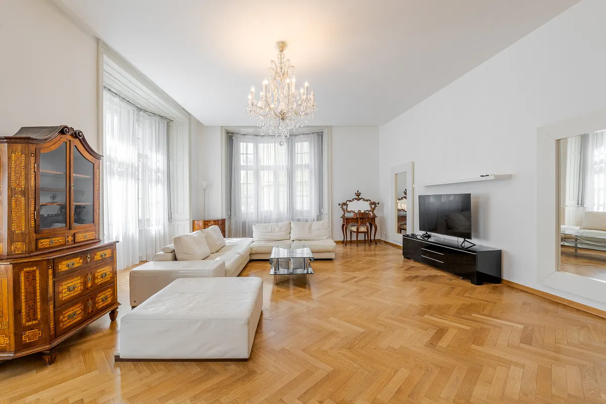 Bright living room with white walls, herringbone wood floors, and a crystal chandelier. A white sectional sofa faces a black TV stand.