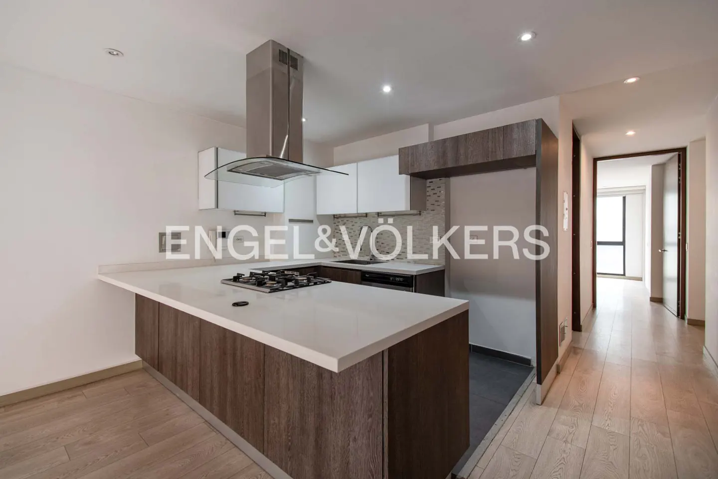 A modern kitchen with white cabinets, a stainless steel range hood, and a large island with a white countertop. A hallway is visible in the background.