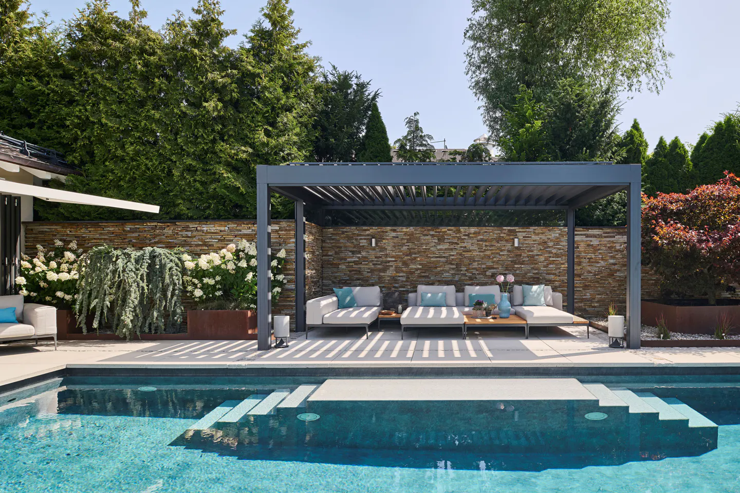 Outdoor patio with a pool, lounge chairs under a pergola, and a stone wall backdrop.