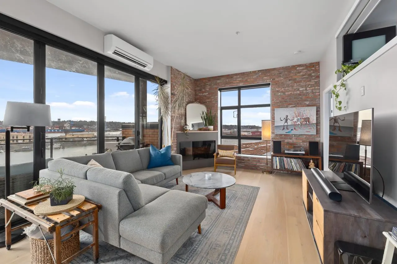 Bright living room with a gray sectional sofa, brick wall, fireplace, and large windows overlooking a cityscape and water.