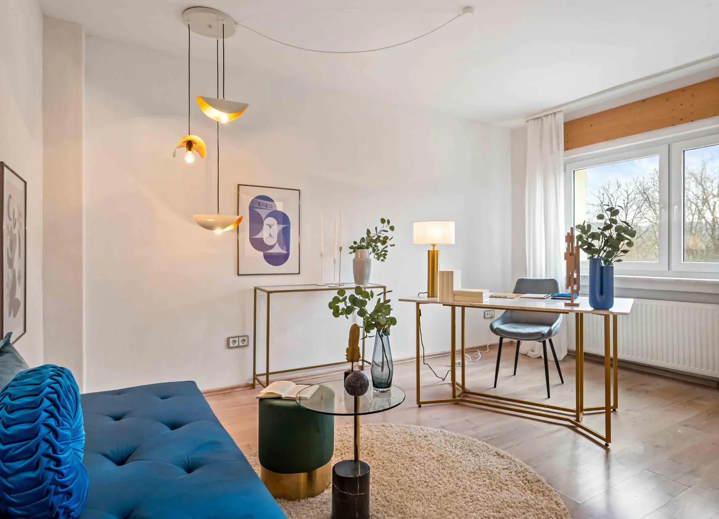 Bright living space with a blue sofa, round rug, and a desk by a window. A modern light fixture hangs above a glass table.