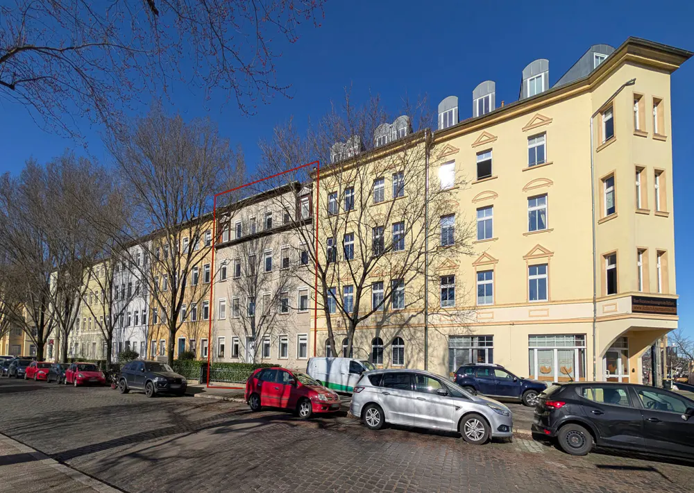 A yellow apartment building with many windows, parked cars, and bare trees under a blue sky.