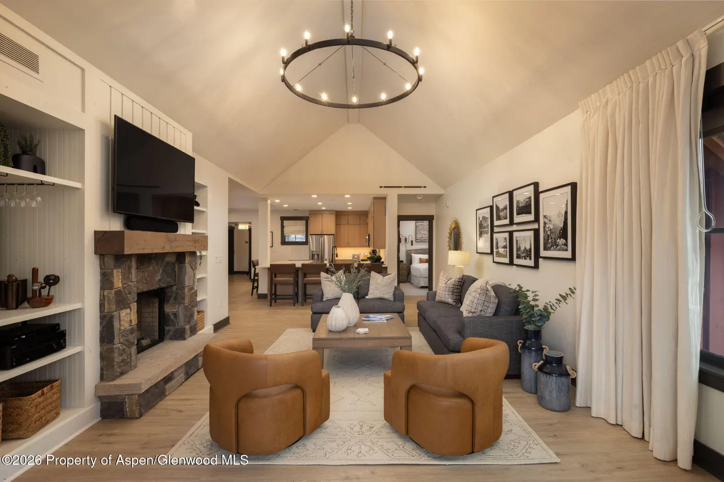Living room with stone fireplace, TV, built-in shelves, two brown chairs, gray sofa, and open kitchen in the background.