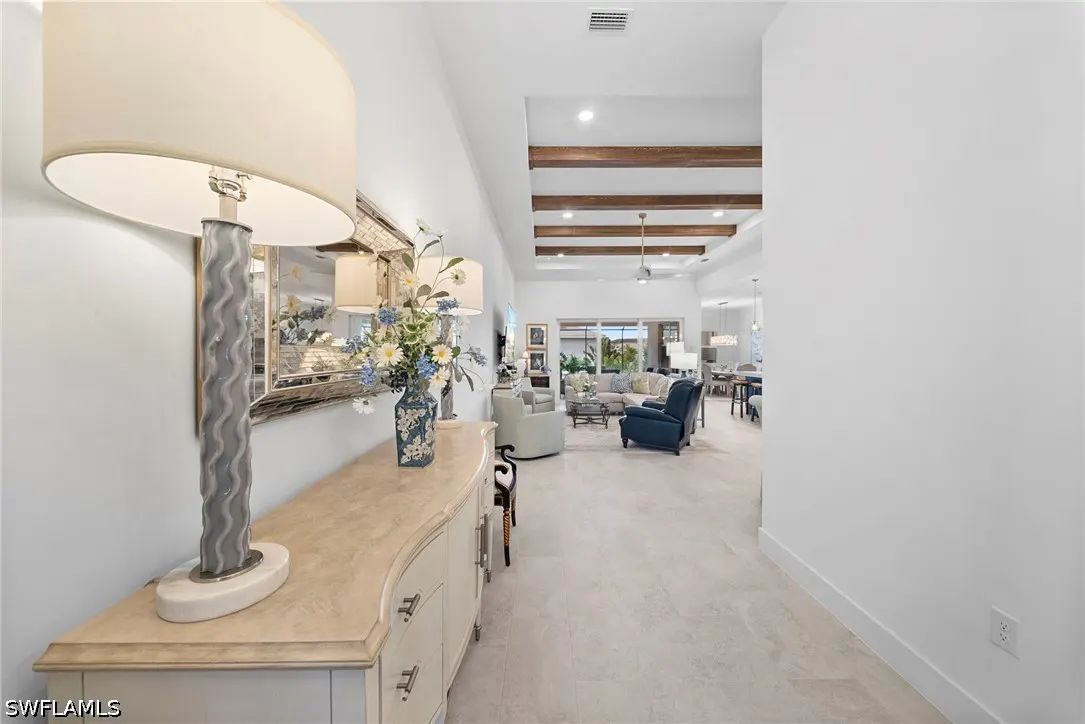 Entryway with a beige console table, lamp, and floral vase, leading to an open-concept living room with wood ceiling beams.