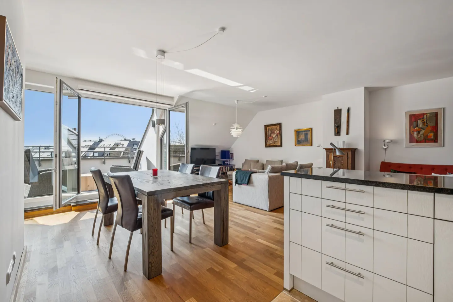Bright, open-plan living space with a dining table, chairs, and balcony doors open to a city view. White walls and wooden floors.