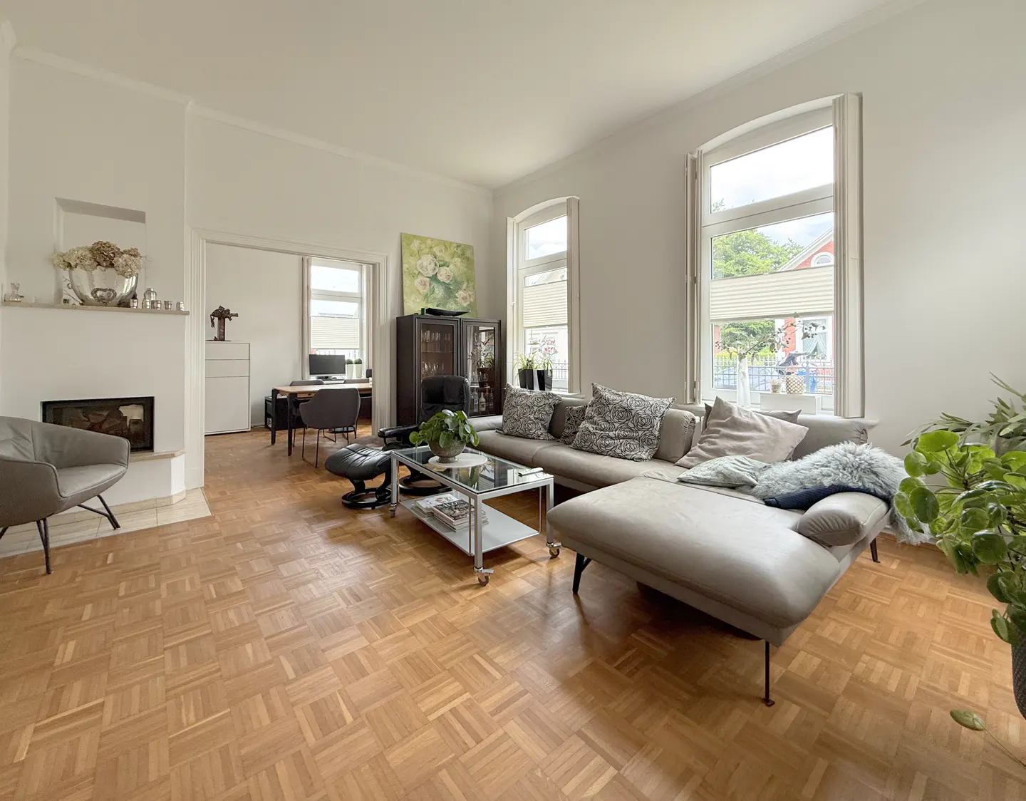 Bright living room with parquet floors, a grey sectional sofa, glass coffee table, fireplace, and a view into an office.