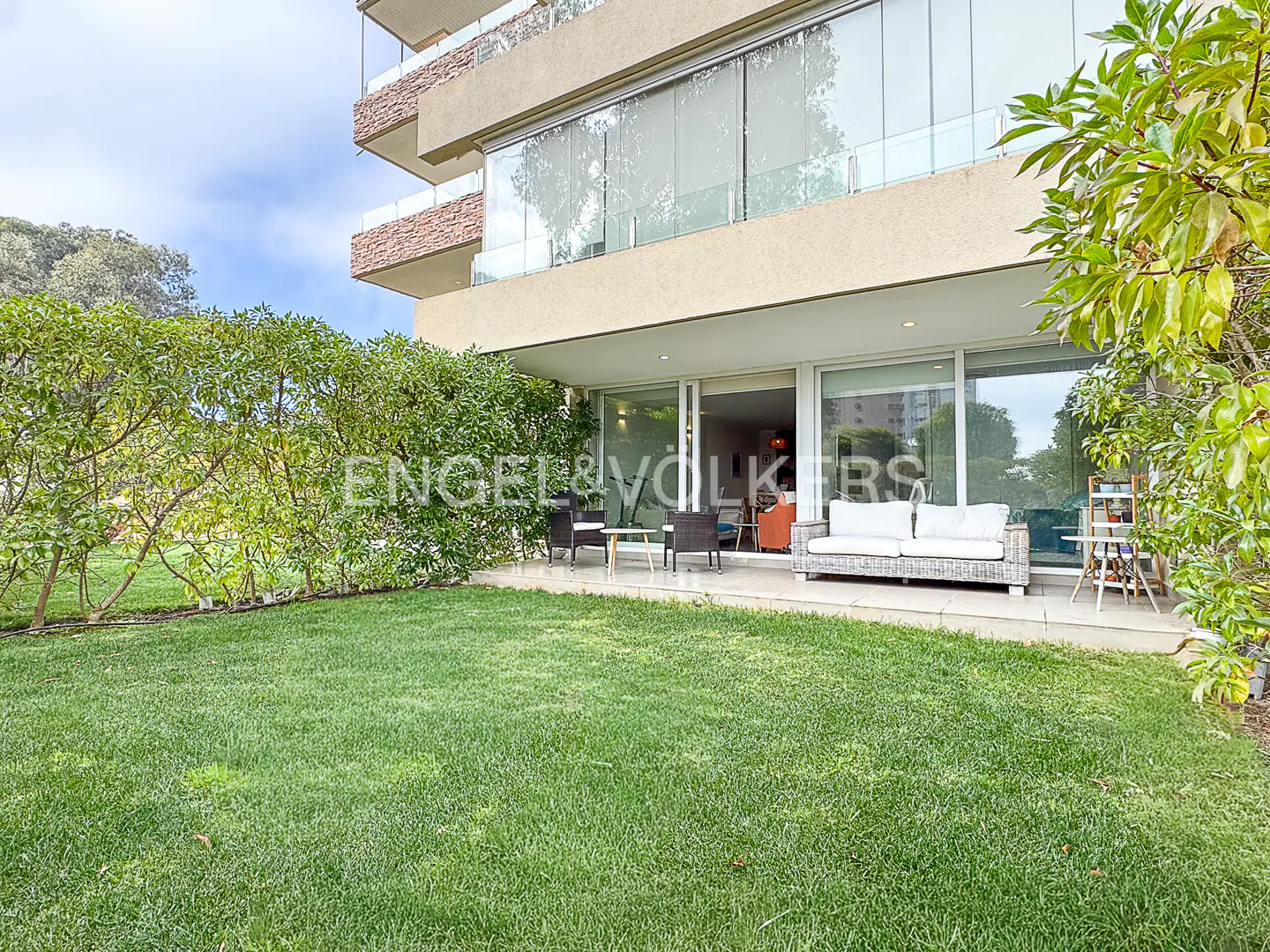 A modern apartment patio with white wicker furniture, overlooking a lush green lawn surrounded by trees.