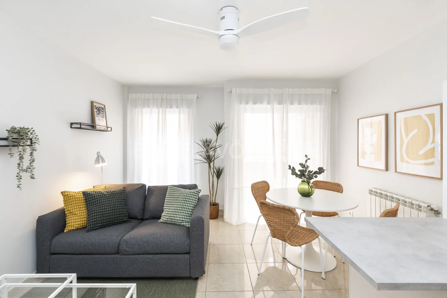 Bright living room with gray sofa, yellow and green pillows, white table with wicker chairs, and white ceiling fan.