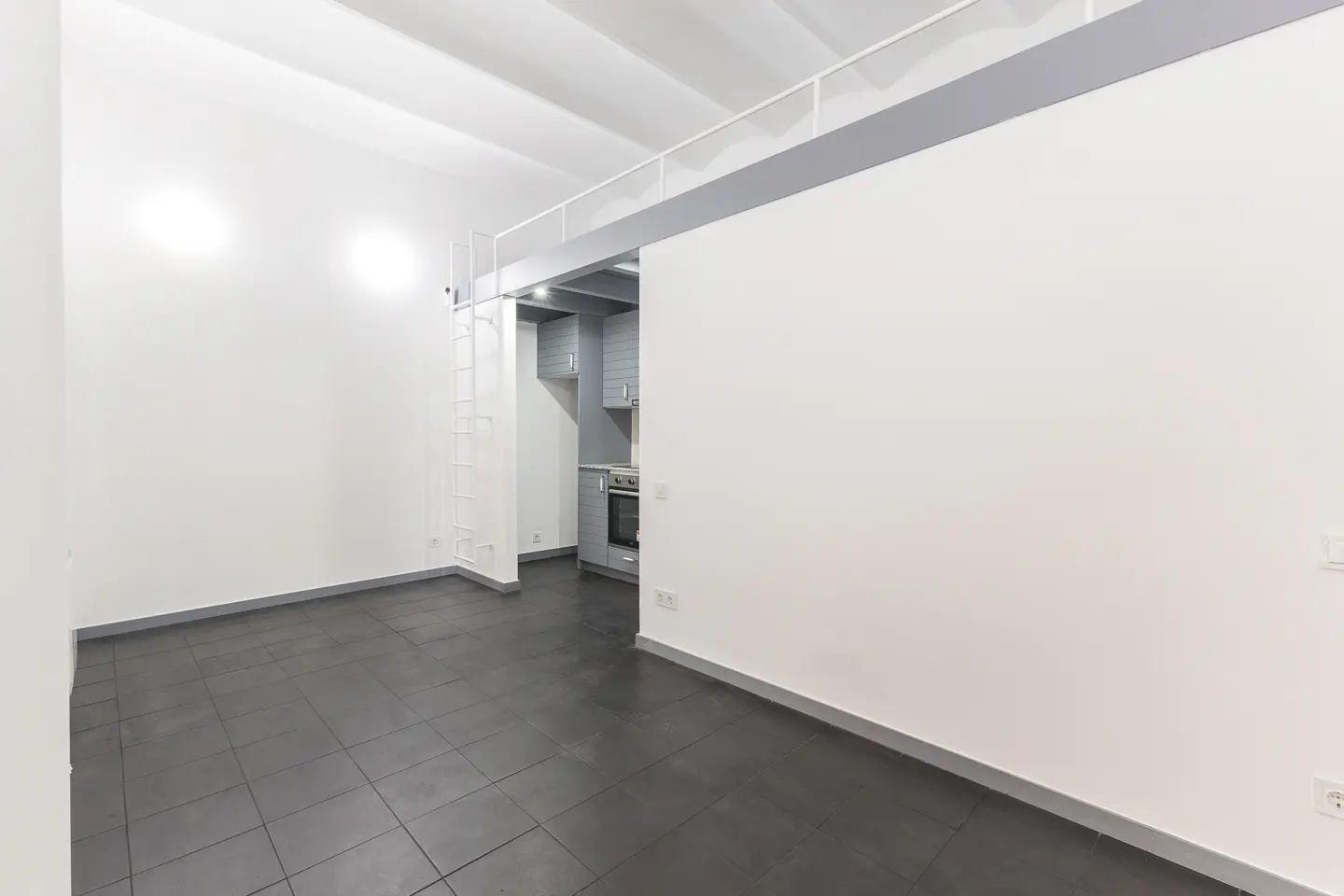 An interior view of a modern apartment with white walls, dark tile floors, and a gray kitchen visible in the background. A white ladder leads to a loft area.