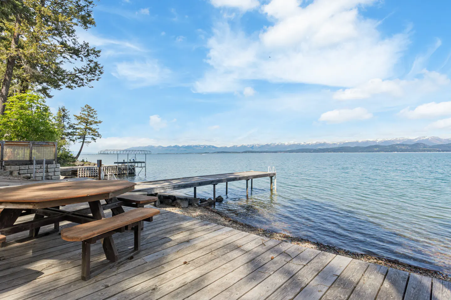 Wooden picnic table on a deck overlooking a lake with a dock, mountains, and blue sky.