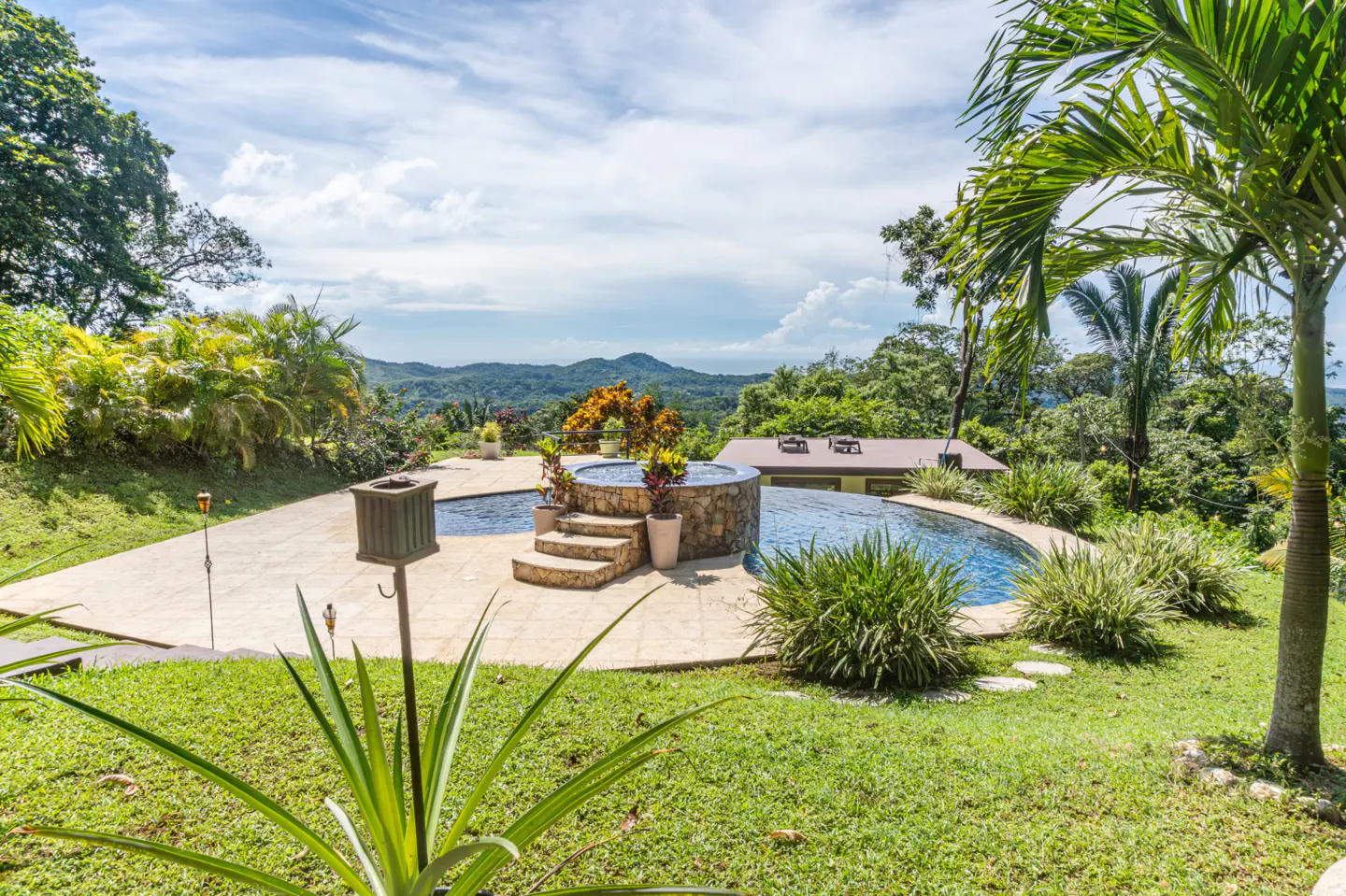 Outdoor pool with stone hot tub and steps, surrounded by lush green grass, palm trees, and a mountain view under a blue sky.