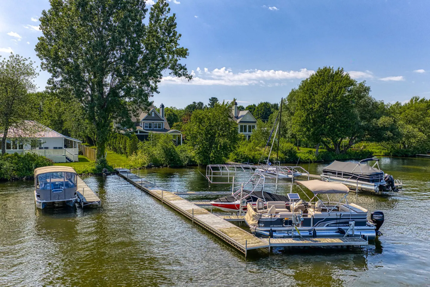 Aerial view of a lake with a dock and several boats, surrounded by trees and houses under a blue sky.