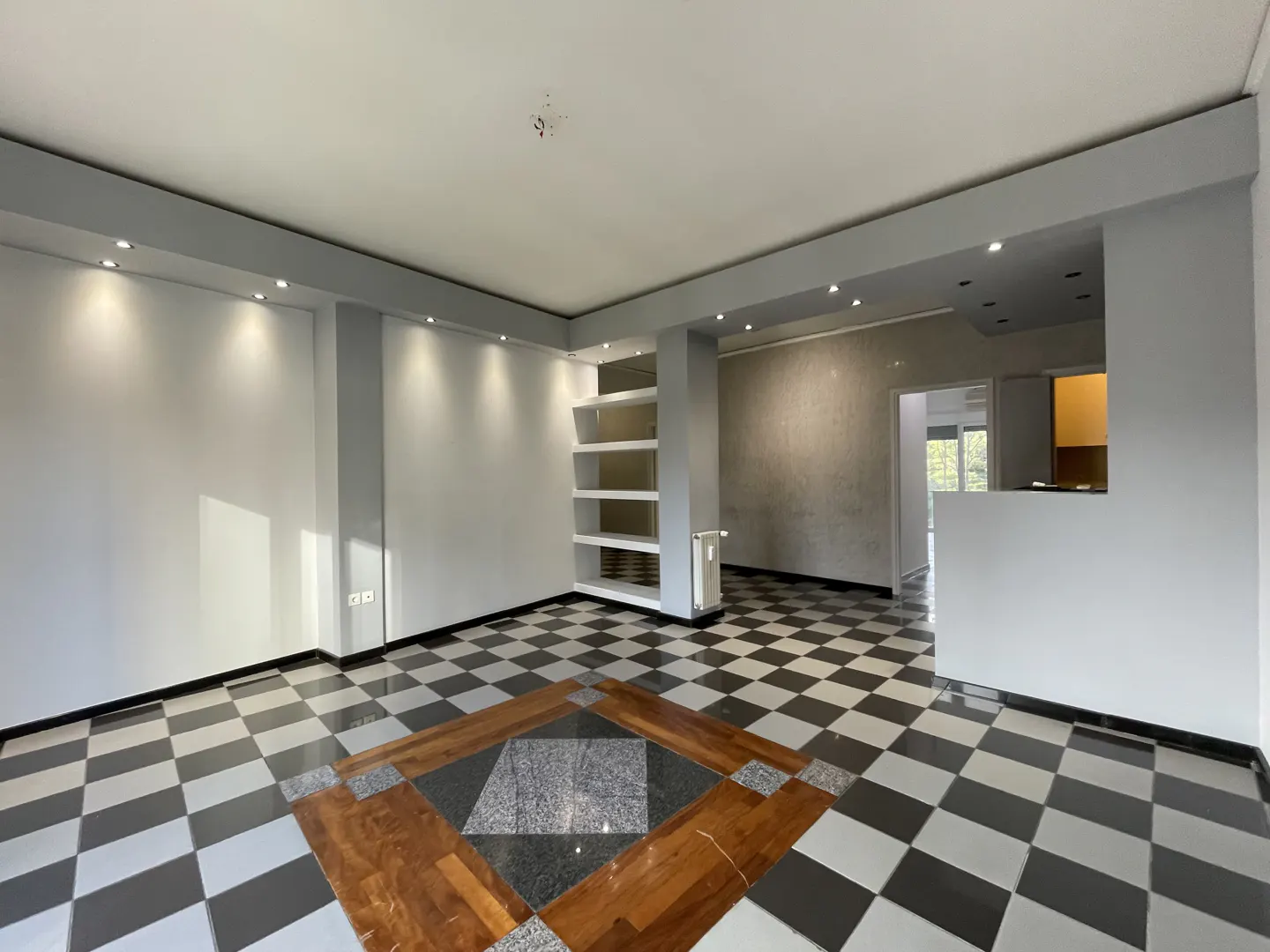 A spacious room with black and white checkered floors, gray walls, and recessed lighting. A white shelving unit is on the left.