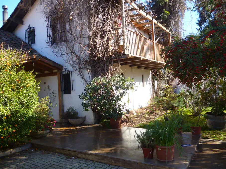 A white house with a wooden balcony is surrounded by lush greenery and potted plants.