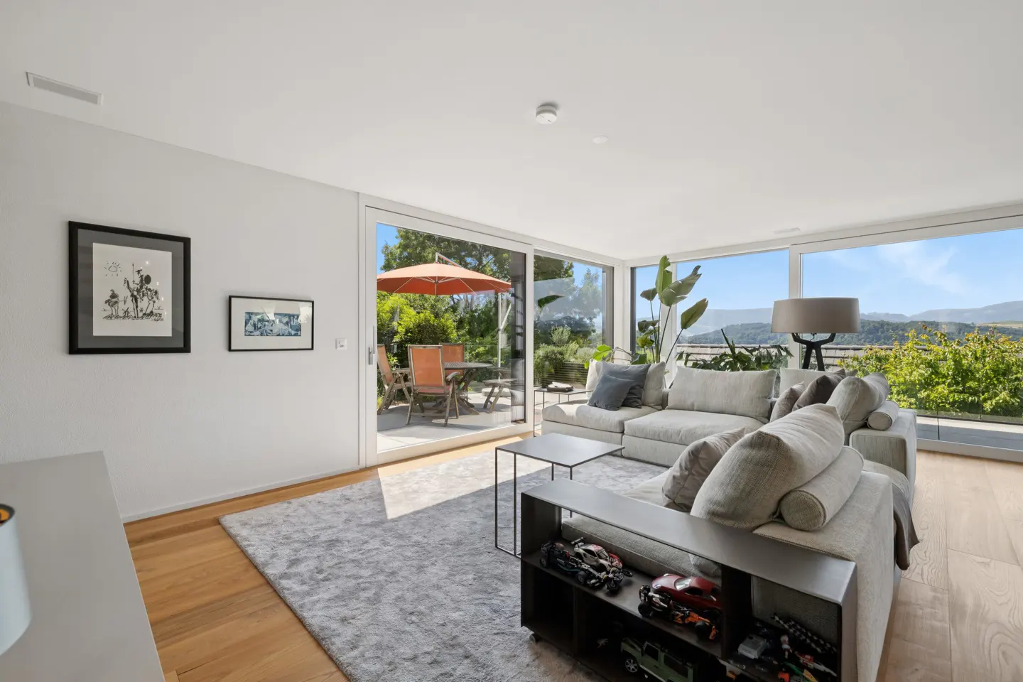 Bright living room with a gray sectional sofa, wood floors, and large windows overlooking a patio with an orange umbrella.