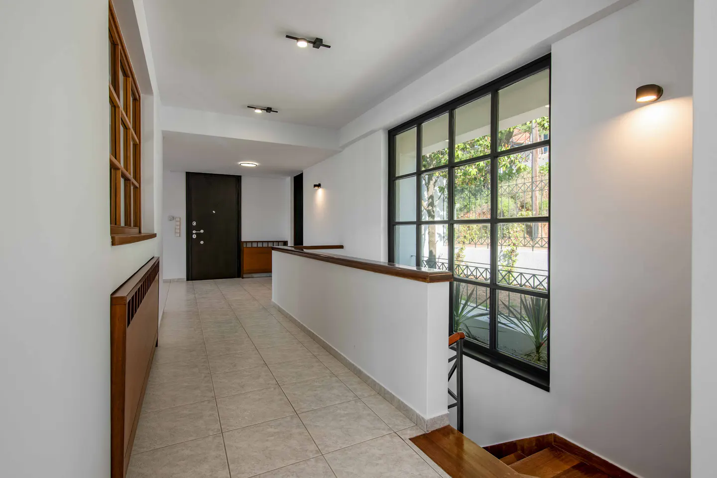 Hallway with white walls, tile floor, and a large black-framed window showing greenery outside. A dark door and wooden radiator are visible.
