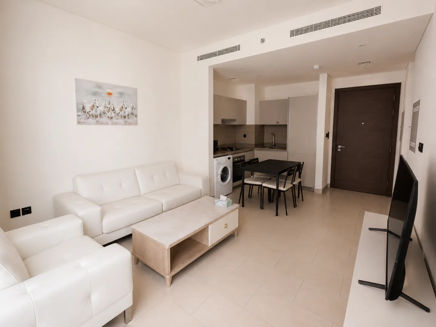 A bright apartment interior with white sofa, coffee table, kitchen, dining table, and TV.