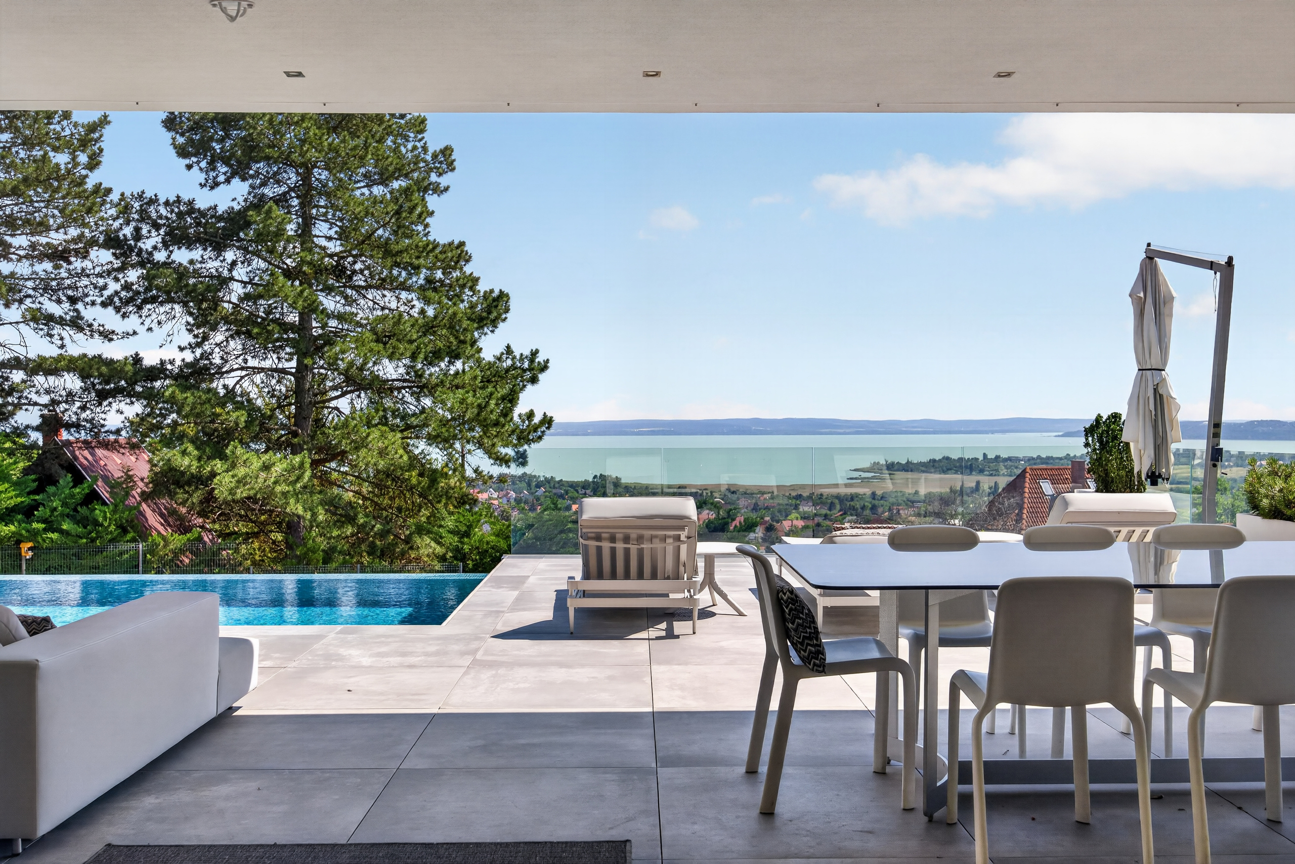 Modern patio with dining set and lounge chairs overlooking an infinity pool, lush trees, and a distant lake view.