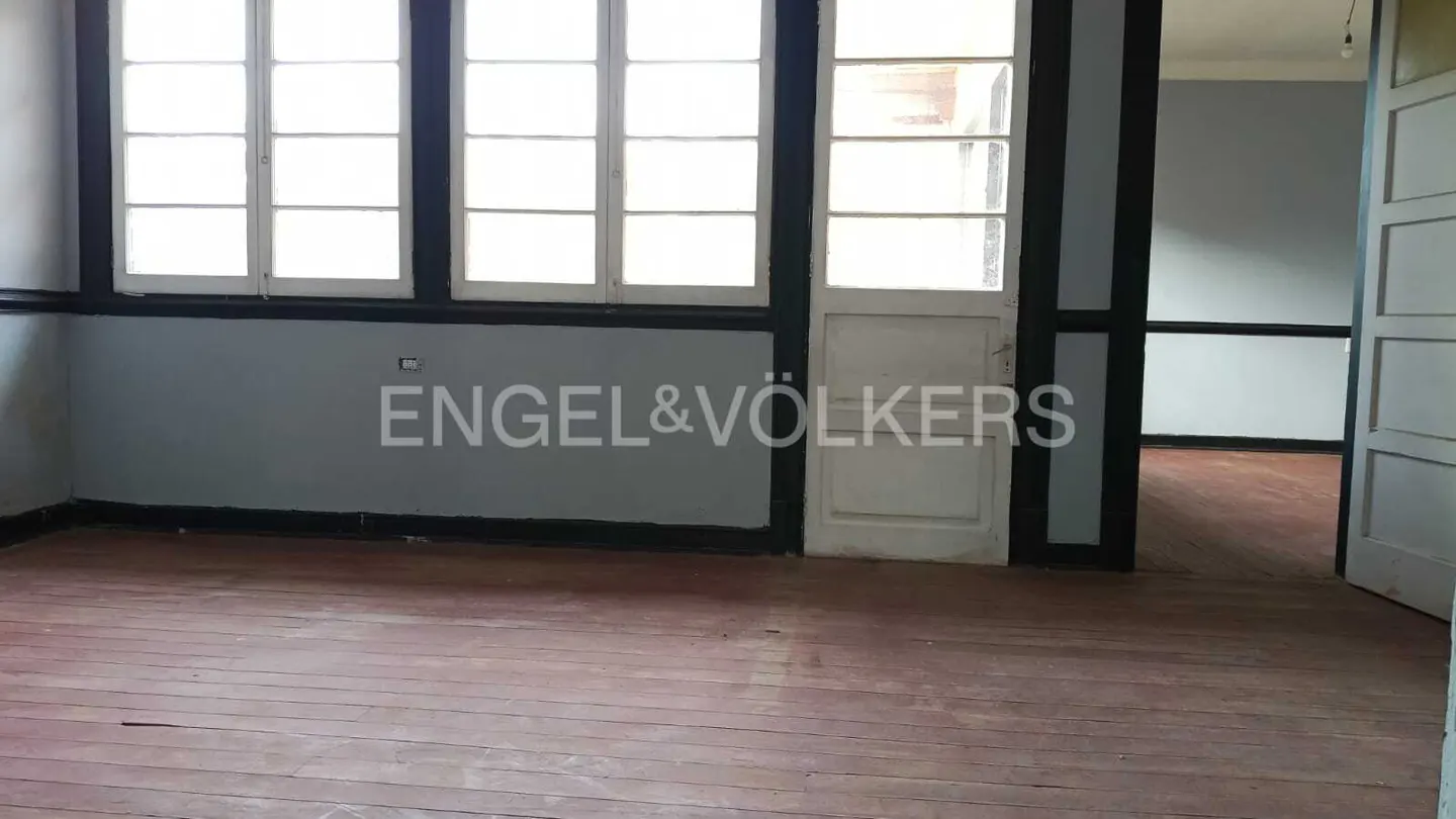 Empty room with hardwood floors, gray walls, and white-framed windows. Engel & Völkers logo is visible on the wall.