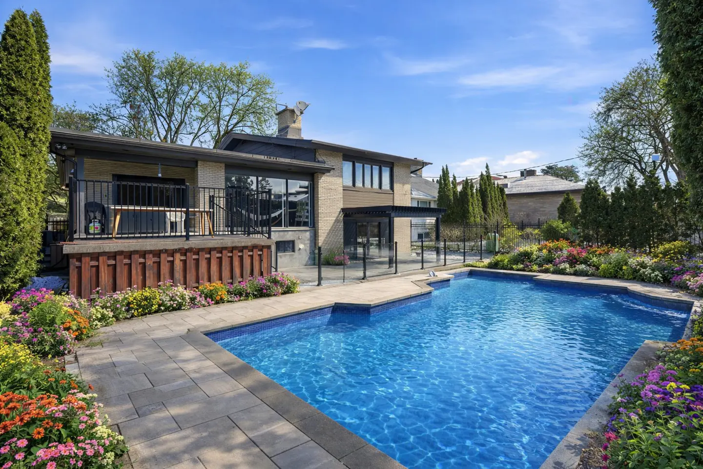 Backyard view of a brick house with a blue tiled pool, patio, and colorful flower beds under a bright blue sky.