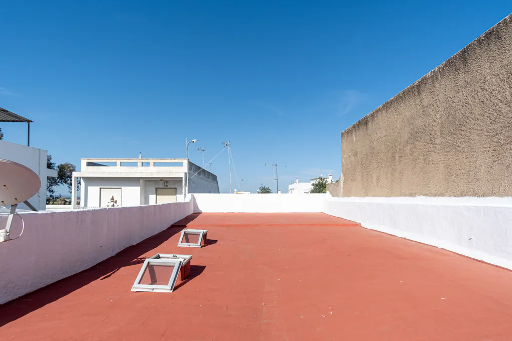 Red rooftop with white parapet walls under a blue sky. Two skylights are visible. Buildings and antennas are in the background.