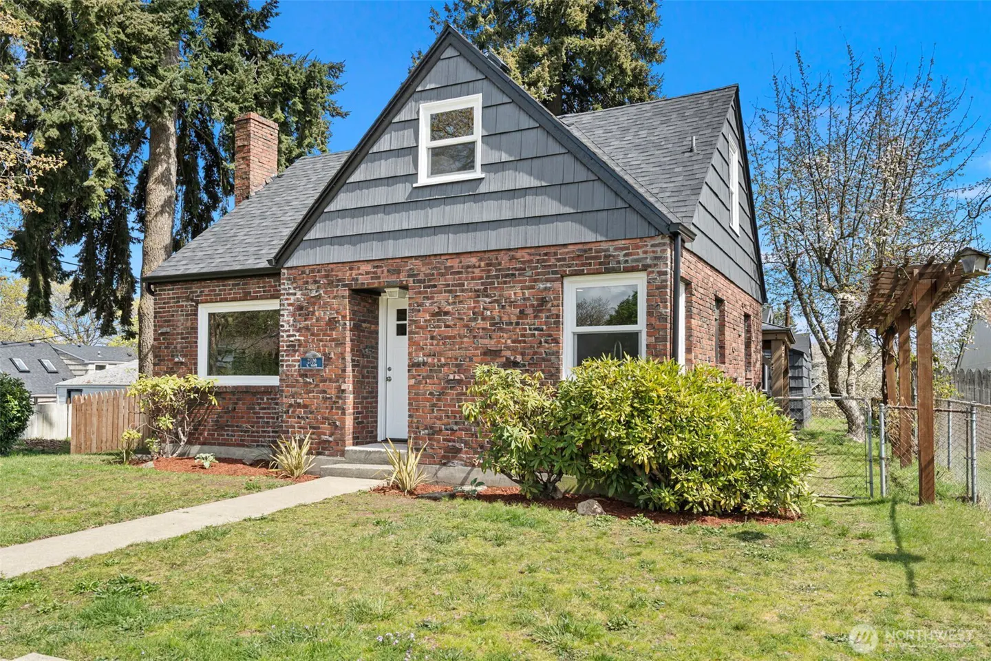 A red brick house with a gray shingle roof and a white front door sits on a green lawn under a blue sky.