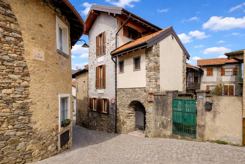 A narrow cobblestone street in a European village with stone buildings and wooden shutters under a blue sky.