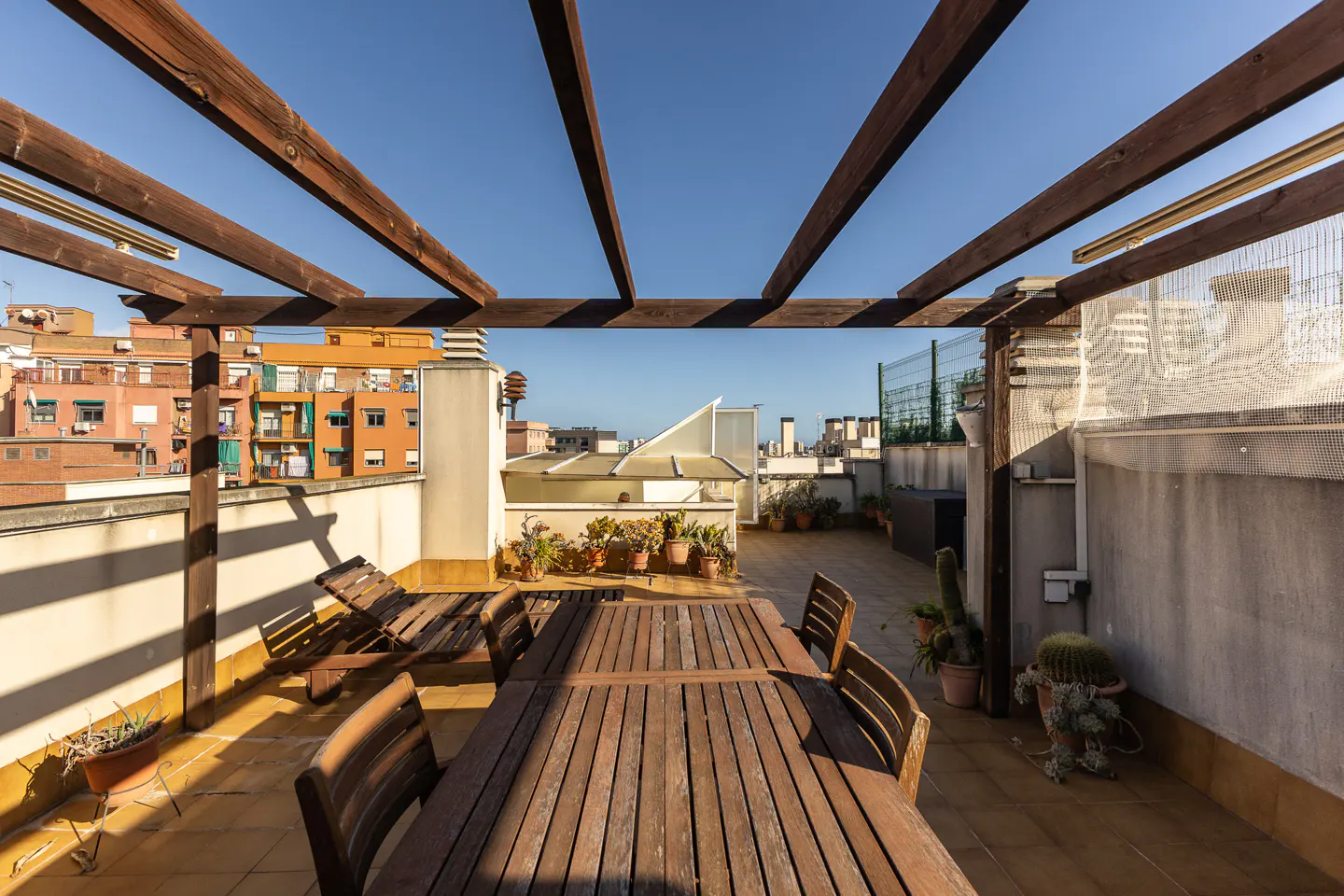 Rooftop terrace with wooden pergola, table, chairs, and lounge chair. Potted plants line the edge, with city buildings in the background under a blue sky.