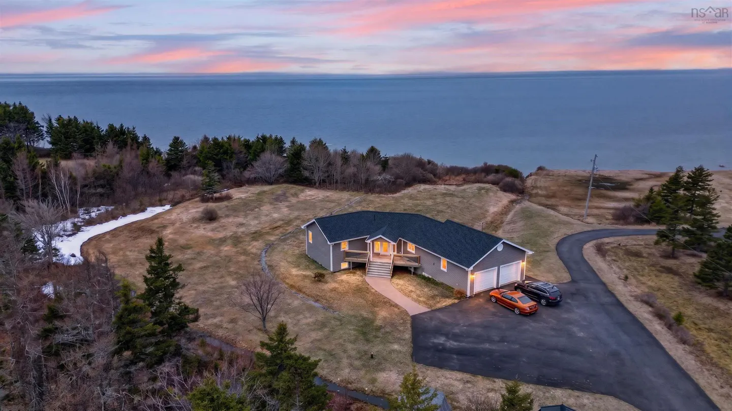 Aerial view of a gray house with a dark roof, a driveway, and two cars, with the ocean in the background.