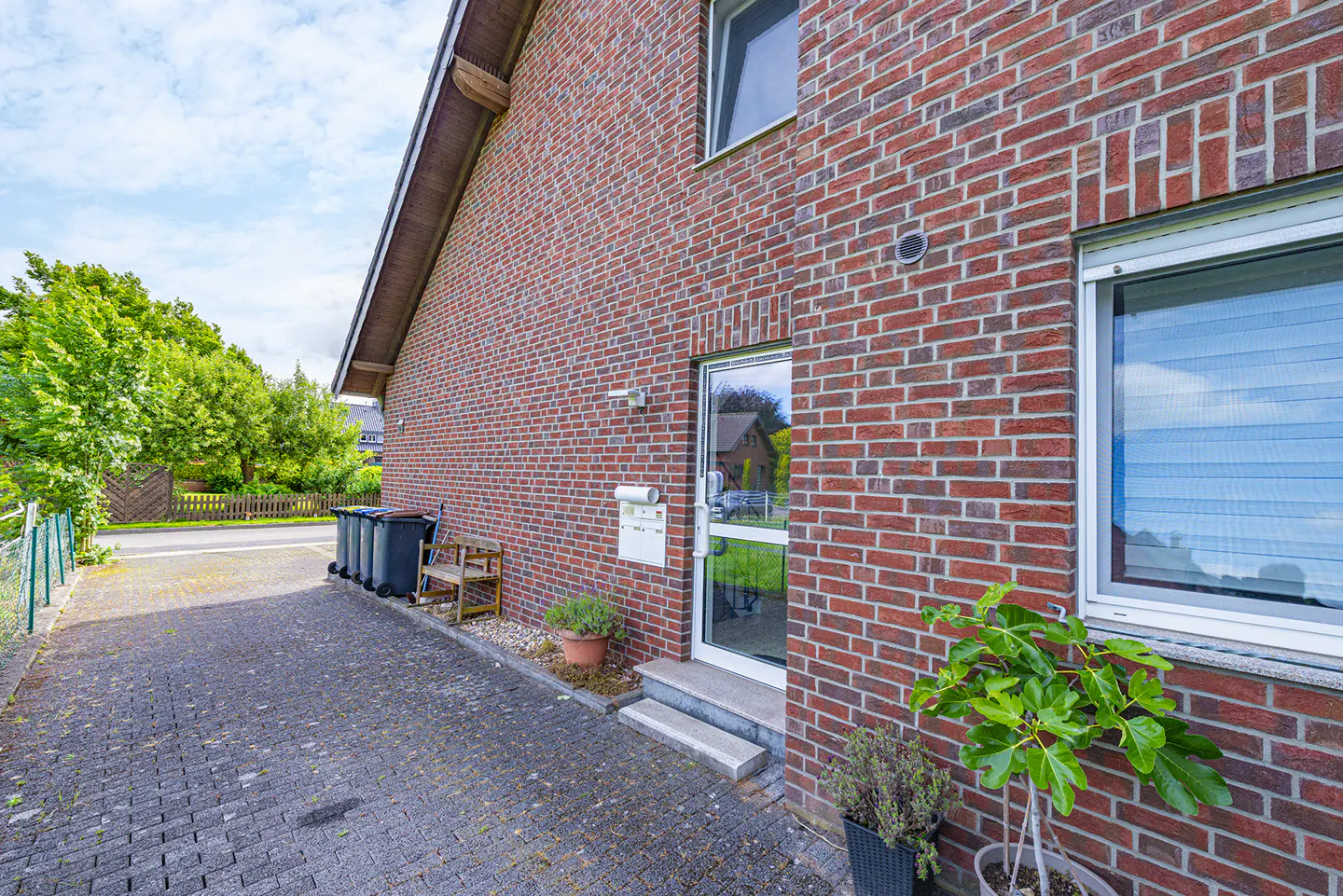 Side view of a red brick house with a glass door, paved driveway, trash bins, and potted plants under a blue sky.