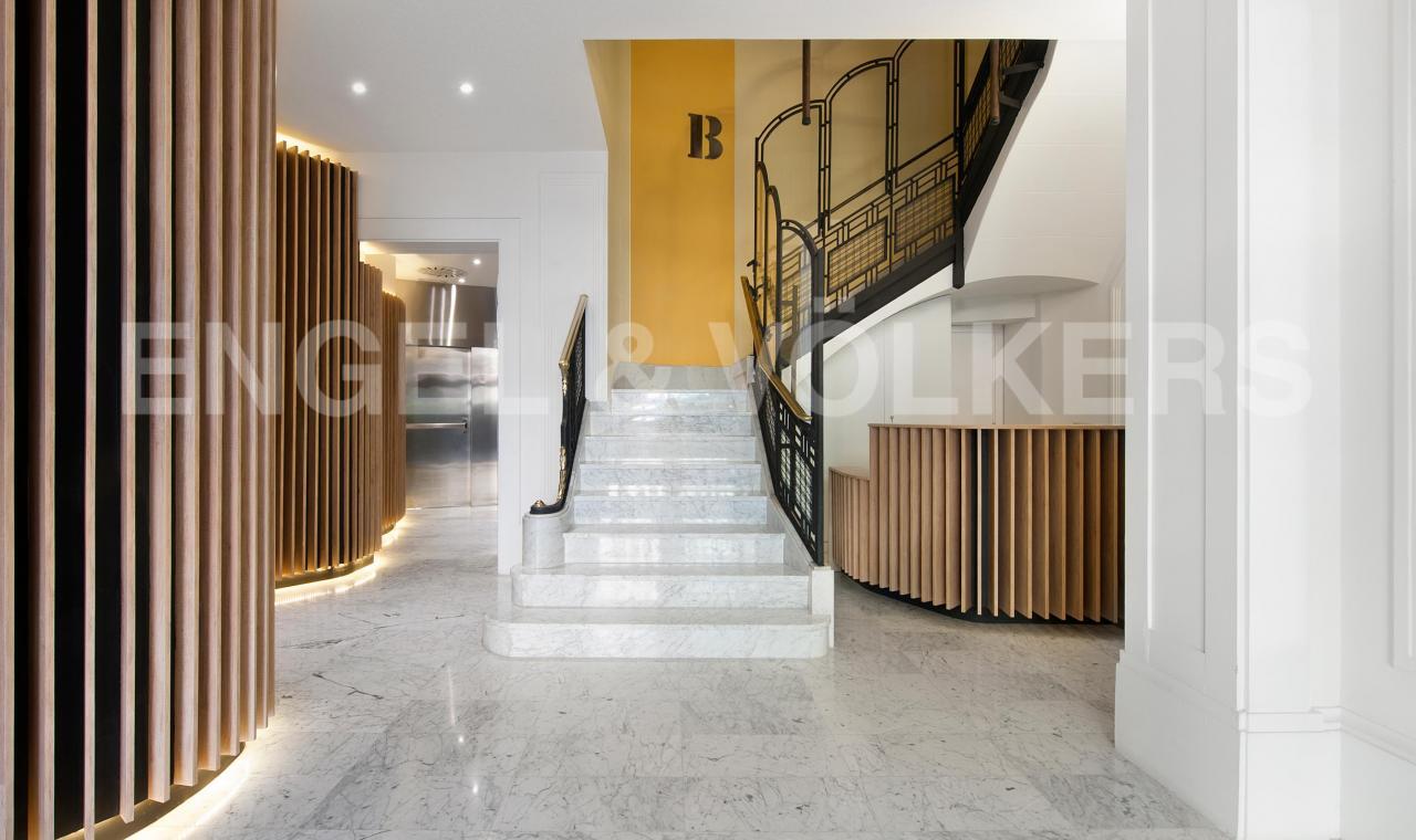 Elegant lobby with marble floors, a staircase with black railings, and wood slat accents. A yellow wall features a large "B".